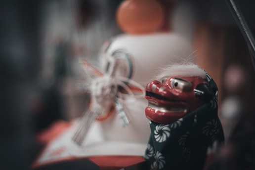 A close-up of a traditional Japanese lion mask, known as a Shishi-mai, with a red face and adorned with a patterned cloth, next to another mask with intricate detailing and ribbons.
