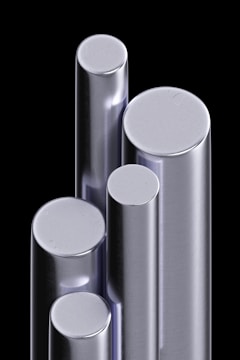 A cluster of shiny, metallic cylindrical bars standing upright against a black background. The bars have varying heights and diameters, reflecting light with a smooth, polished surface.
