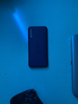 A flat lay image featuring a black smartphone with a leather case, a partial view of a laptop, and part of a gaming controller against a blue illuminated surface.