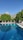 A tranquil outdoor swimming pool surrounded by sun loungers and white umbrellas, set against a backdrop of lush green trees and majestic mountains in the distance.