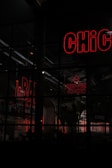 A scene depicting a dimly lit restaurant or bar area with neon signs. The words 'BAR' and 'PERi-PERi CHICKEN' are illuminated in red neon, and part of a 'CHiC' sign is visible. The establishment appears closed as indicated by a sign. Shelves with bottles are visible in the background, along with some interior plant decor.