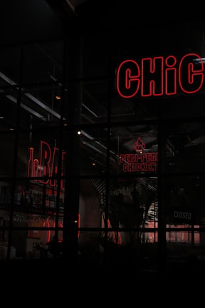 A scene depicting a dimly lit restaurant or bar area with neon signs. The words 'BAR' and 'PERi-PERi CHICKEN' are illuminated in red neon, and part of a 'CHiC' sign is visible. The establishment appears closed as indicated by a sign. Shelves with bottles are visible in the background, along with some interior plant decor.