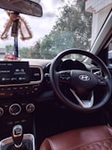Inside view of a car focusing on the steering wheel with a visible car brand logo. The dashboard shows a display with options like media and settings. Hanging from the rearview mirror is a decorative item with a 'AREON' tag. Through the windshield, trees and part of a building are visible, indicating the car is parked.