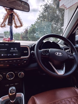 Inside view of a car focusing on the steering wheel with a visible car brand logo. The dashboard shows a display with options like media and settings. Hanging from the rearview mirror is a decorative item with a 'AREON' tag. Through the windshield, trees and part of a building are visible, indicating the car is parked.