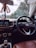 Inside view of a car focusing on the steering wheel with a visible car brand logo. The dashboard shows a display with options like media and settings. Hanging from the rearview mirror is a decorative item with a 'AREON' tag. Through the windshield, trees and part of a building are visible, indicating the car is parked.