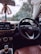 Inside view of a car focusing on the steering wheel with a visible car brand logo. The dashboard shows a display with options like media and settings. Hanging from the rearview mirror is a decorative item with a 'AREON' tag. Through the windshield, trees and part of a building are visible, indicating the car is parked.