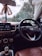 Inside view of a car focusing on the steering wheel with a visible car brand logo. The dashboard shows a display with options like media and settings. Hanging from the rearview mirror is a decorative item with a 'AREON' tag. Through the windshield, trees and part of a building are visible, indicating the car is parked.