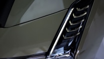 A close-up of a modern car headlight with a sleek, angular design. The light housing features a series of horizontal slats and a vertical strip of LED lights.