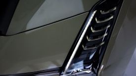 A close-up of a modern car headlight with a sleek, angular design. The light housing features a series of horizontal slats and a vertical strip of LED lights.