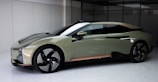 A sleek, modern car with an aerodynamic design and matte finish is parked indoors. The car features a two-toned color scheme with a green and black body, large black wheels, and a subtle hint of copper near the base. The setting appears clean and minimalist, accentuating the car’s futuristic aesthetic.