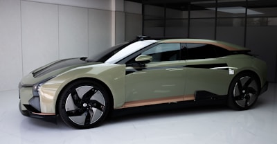 A sleek, modern car with an aerodynamic design and matte finish is parked indoors. The car features a two-toned color scheme with a green and black body, large black wheels, and a subtle hint of copper near the base. The setting appears clean and minimalist, accentuating the car’s futuristic aesthetic.