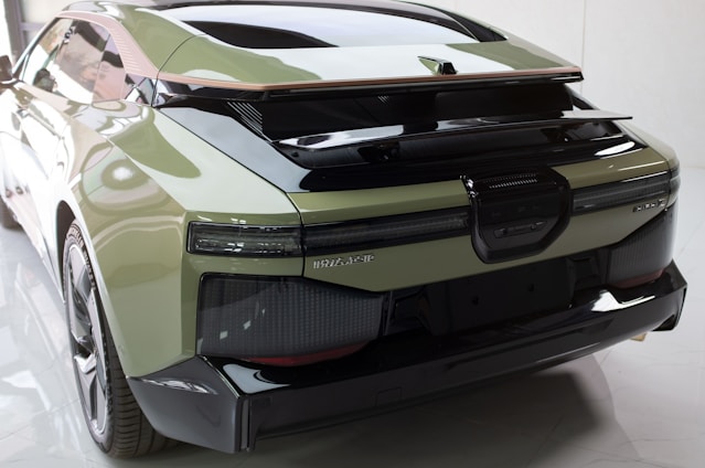 A sleek, modern car with a glossy finish, showcasing an aerodynamic design. The rear view highlights its broad tail lights, reflector features, and a prominent spoiler. Smooth lines and curves add to the futuristic appeal, with the car's mint green color contrasting the black accents.