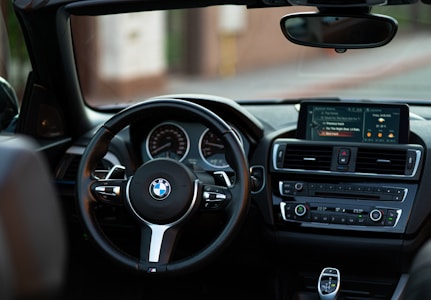 The interior of a BMW car is shown, featuring a sleek steering wheel with the BMW logo prominently displayed in the center. The dashboard displays a digital screen showing music selections and car settings. The controls are detailed with buttons and dials for navigation, air conditioning, and radio functionality.