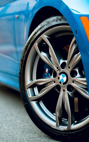 Close-up view of a modern car wheel featuring a five-spoke alloy rim with a BMW logo. The tire appears well-worn, and the body of the car is painted in a vibrant blue. The scene is set on a paved surface, and the image exudes a sense of speed and craftsmanship.