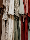 A collection of various dresses is lined up, showcasing a range of patterns and fabrics. The dresses include floral prints, gingham patterns, and solid colors, hung closely together.