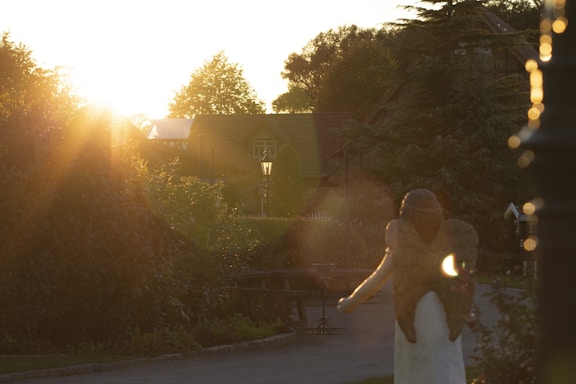 A serene garden scene during sunset with a warm, soft glow filling the area. A statue of an angel with outstretched wings stands on a pathway. The sun's rays create a lens flare effect and highlight the lush greenery, with a rustic house in the background. A vintage-style street lamp can be seen near the center of the image.