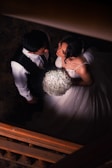 A man and a woman are standing close together in an intimate setting, possibly on a staircase. The woman is wearing a white gown and holding a bouquet of small white flowers. The man is dressed in formal attire, wearing a black vest and white shirt.
