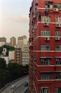 A tall red residential building with several air conditioning units fixed to its exterior, and a few individuals visibly performing maintenance work or cleaning on the windows. A curved road runs parallel to the building, with a couple of cars driving by. In the distance, other high-rise buildings are visible, along with trees providing some greenery to the urban landscape.