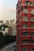 A tall red residential building with several air conditioning units fixed to its exterior, and a few individuals visibly performing maintenance work or cleaning on the windows. A curved road runs parallel to the building, with a couple of cars driving by. In the distance, other high-rise buildings are visible, along with trees providing some greenery to the urban landscape.