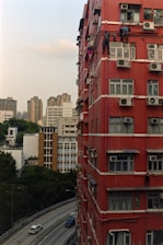 A tall red residential building with several air conditioning units fixed to its exterior, and a few individuals visibly performing maintenance work or cleaning on the windows. A curved road runs parallel to the building, with a couple of cars driving by. In the distance, other high-rise buildings are visible, along with trees providing some greenery to the urban landscape.
