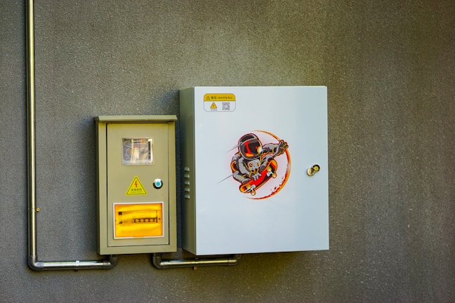 An electrical control panel is mounted on a textured gray wall. The panel consists of two units: one is a smaller beige box with a window displaying internal components, and a warning sign with Chinese characters and a lightning bolt symbol. The other box is larger, white, and features an illustration of a cartoon character wearing a helmet and armor, riding a skateboard in a dynamic pose.
