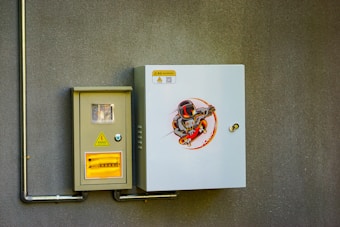 An electrical control panel is mounted on a textured gray wall. The panel consists of two units: one is a smaller beige box with a window displaying internal components, and a warning sign with Chinese characters and a lightning bolt symbol. The other box is larger, white, and features an illustration of a cartoon character wearing a helmet and armor, riding a skateboard in a dynamic pose.