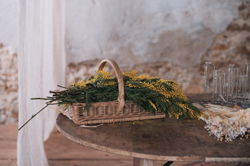 A wicker basket filled with green foliage and yellow flowers sits on a wooden round table. The background features a textured, partially damaged wall with a mix of plaster and exposed stone. Next to the basket on the table, there are glass candle holders and a bunch of dried flowers. White sheer fabric is draped to the left side of the scene.