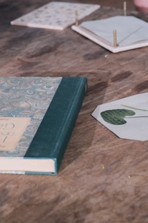 A hardcover book with a decorative cover and green spine lies on a wooden table. Nearby, there are a few coasters, one with a leaf imprint and others with small pins protruding from them. The surface of the table has a rustic appearance.