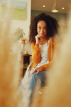 A woman with curly hair wearing a striped shirt and an orange cardigan is sitting thoughtfully on a chair. She appears relaxed and content in a warmly lit room with decor that includes plants, a painting on the wall, and a modern hanging lamp.