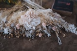 A beautifully arranged bouquet of dried flowers, featuring a large pale pink rose at the center surrounded by various types of dried flowers and greenery. The arrangement includes small seed pods, delicate white and cream-colored flowers, and other textured botanical elements. It rests against a textured brown surface, adding a rustic and vintage ambiance.