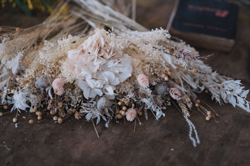 A beautifully arranged bouquet of dried flowers, featuring a large pale pink rose at the center surrounded by various types of dried flowers and greenery. The arrangement includes small seed pods, delicate white and cream-colored flowers, and other textured botanical elements. It rests against a textured brown surface, adding a rustic and vintage ambiance.