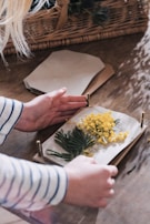 Hands assembling a botanical press with various green foliage and small yellow flowers placed between paper sheets. The setup is on a wooden surface, and a wicker basket is visible in the background.