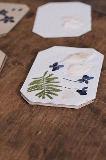 A wooden surface features rectangular coasters decorated with botanical elements. One coaster prominently displays a green fern, blue flowers, and white petals artistically arranged. Other coasters are partially visible with similar natural motifs.