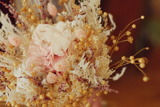 A bouquet of dried flowers featuring a mix of delicate textures. Soft pink petals are surrounded by cream and beige elements, including seed pods and feathery foliage. The arrangement displays a harmonious blend of natural forms.