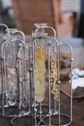 A group of glass test tubes are arranged in metal holders. Some of the test tubes contain clear liquid and are decorated with small yellow flowers. The background is a soft blur of neutral colors.