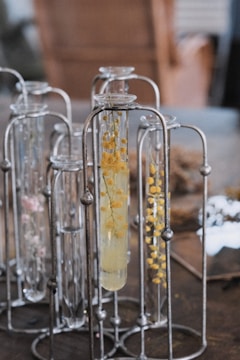 A group of glass test tubes are arranged in metal holders. Some of the test tubes contain clear liquid and are decorated with small yellow flowers. The background is a soft blur of neutral colors.