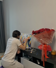 A person wearing a white t-shirt and gray pants is kneeling and painting on a vibrant abstract canvas placed on a black shelf. Beside the painting, there is a large bouquet of red roses wrapped in cellophane. Small paint containers and a brush are seen on the shelf. A dark curtain is visible in the background.