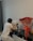 A person wearing a white t-shirt and gray pants is kneeling and painting on a vibrant abstract canvas placed on a black shelf. Beside the painting, there is a large bouquet of red roses wrapped in cellophane. Small paint containers and a brush are seen on the shelf. A dark curtain is visible in the background.