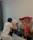 A person wearing a white t-shirt and gray pants is kneeling and painting on a vibrant abstract canvas placed on a black shelf. Beside the painting, there is a large bouquet of red roses wrapped in cellophane. Small paint containers and a brush are seen on the shelf. A dark curtain is visible in the background.