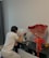 A person wearing a white t-shirt and gray pants is kneeling and painting on a vibrant abstract canvas placed on a black shelf. Beside the painting, there is a large bouquet of red roses wrapped in cellophane. Small paint containers and a brush are seen on the shelf. A dark curtain is visible in the background.