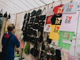 A person wearing a blue and red jacket stands in front of a market stall displaying various items. The stall is filled with black bags and colorful t-shirts, which are hanging from racks. The t-shirts predominantly feature the 'I ❤️ HK' slogan in different colors. There is also a green shirt with a Hong Kong design and a price tag indicating a sale.