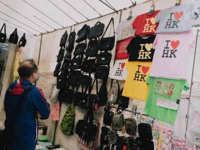 A person wearing a blue and red jacket stands in front of a market stall displaying various items. The stall is filled with black bags and colorful t-shirts, which are hanging from racks. The t-shirts predominantly feature the 'I ❤️ HK' slogan in different colors. There is also a green shirt with a Hong Kong design and a price tag indicating a sale.