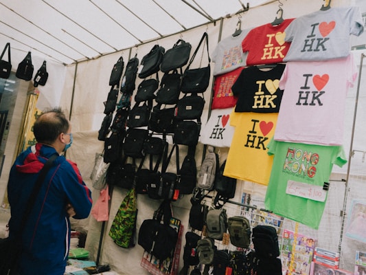A person wearing a blue and red jacket stands in front of a market stall displaying various items. The stall is filled with black bags and colorful t-shirts, which are hanging from racks. The t-shirts predominantly feature the 'I ❤️ HK' slogan in different colors. There is also a green shirt with a Hong Kong design and a price tag indicating a sale.