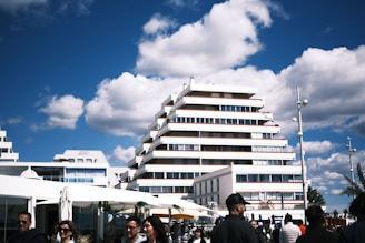 A modern, tiered building stands against a bright blue sky with scattered fluffy clouds. People walk in the foreground, and there are white umbrellas and shops nearby. The architecture is characterized by clean, straight lines and large windows.