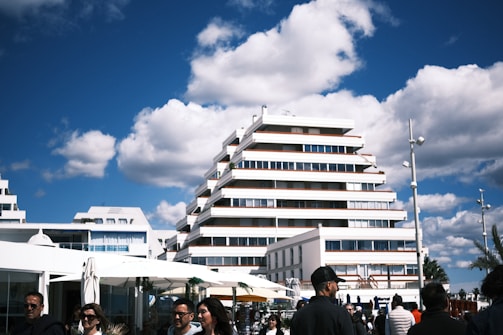 A modern, tiered building stands against a bright blue sky with scattered fluffy clouds. People walk in the foreground, and there are white umbrellas and shops nearby. The architecture is characterized by clean, straight lines and large windows.
