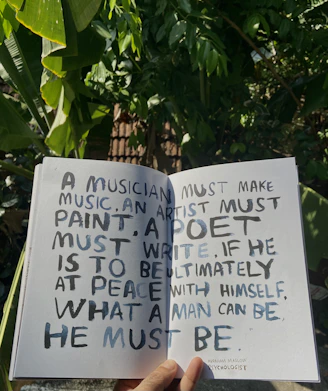A hand holds open a book against a backdrop of lush green leaves, revealing a page with handwritten text. The text discusses the necessity for creatives to engage in their craft to achieve personal fulfillment, with a quote from Abraham Maslow.