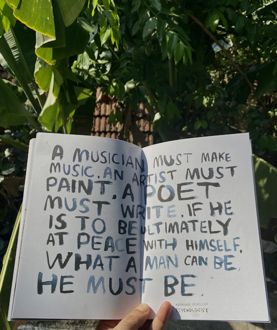 A hand holds open a book against a backdrop of lush green leaves, revealing a page with handwritten text. The text discusses the necessity for creatives to engage in their craft to achieve personal fulfillment, with a quote from Abraham Maslow.