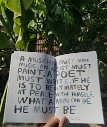 A hand holds open a book against a backdrop of lush green leaves, revealing a page with handwritten text. The text discusses the necessity for creatives to engage in their craft to achieve personal fulfillment, with a quote from Abraham Maslow.