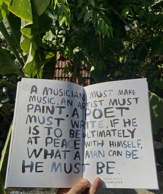 A hand holds open a book against a backdrop of lush green leaves, revealing a page with handwritten text. The text discusses the necessity for creatives to engage in their craft to achieve personal fulfillment, with a quote from Abraham Maslow.