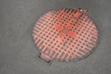 A red manhole cover with a grid pattern lies on a concrete surface. The cover has square indents and a small circular feature at the center. Some wear and chipping are visible, giving it a weathered appearance. It’s positioned slightly off-center with a metal handle visible at the edge.